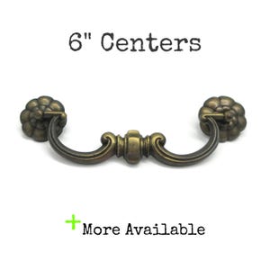 May include: A brass-toned metal drawer pull with a curved handle and two circular, flower-shaped mounting plates. The text 6" Centers is printed above the handle, and the text + More Available is printed below the handle on a white background.