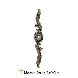 May include: A decorative metal accent piece with a scroll design in an antique copper finish. The piece has a square hole in the center.