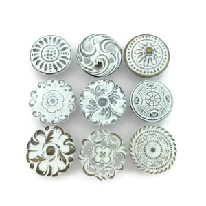 May include: Nine decorative cabinet knobs with various embossed designs. The knobs are round and feature intricate patterns in white and antique gold finishes. The designs include floral, swirl, and geometric motifs.