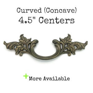 May include: An antique brass-toned drawer pull with a curved, concave design. The pull features ornate, floral-inspired detailing on each end and a curved handle. The text reads "Curved (Concave) 4.5" Centers" and "More Available".