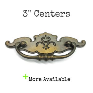 May include: A brass-toned metal drawer pull with a decorative backplate featuring engraved scrollwork and a swinging handle. The text 3" Centers is printed above the hardware, and the text + More Available is printed below the hardware.