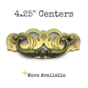 May include: Antique brass drawer pull with a decorative design. The pull has a curved handle and a backplate with floral and geometric details. The text "4.25" Centers" is above the pull. The text "More Available" is below the pull.