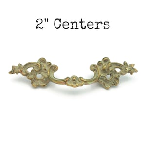 May include: Ornate brass drawer pull with a 2-inch center-to-center measurement. The pull features a curved handle and decorative floral accents on each end. The text "2" Centers" is printed above the pull.
