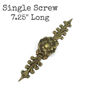 May include: An antique brass-toned metal drawer pull featuring an elongated, ornate backplate with scrolling leaf details and a central circular floral knob. The text "Single Screw 7.25" Long" is printed above the hardware piece.