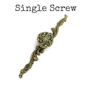 May include: A single antique brass screw with a floral center and ornate, curved arms. The text "Single Screw" is printed above the screw. The screw is designed for furniture or decorative applications.