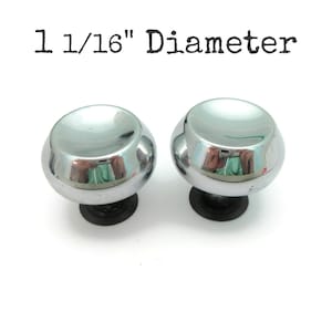 May include: Two shiny chrome cabinet knobs with black bases. The knobs have a rounded, mushroom-like shape and reflect the surrounding environment. The text "1 1/16" Diameter" is above the knobs.