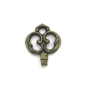 May include: An antique-style key charm with an ornate design. The key is made of a bronze-colored metal and features a decorative top with a series of swirls and curves. The key's stem is rectangular and plain.
