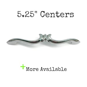 May include: A silver-toned metal drawer pull with a curved, elongated shape and a central decorative element. The text 5.25" Centers is printed above the handle, and the text + More Available is printed below the handle in green.