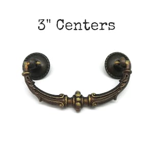 May include: A dark bronze-toned metal drawer pull with a curved handle and two circular mounting plates. The handle features decorative ridges and scrollwork details. The text 3" Centers is printed above the hardware, indicating the distance between the two mounting points in inches.