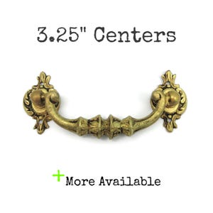May include: A brass-toned metal drawer pull with an ornate, curved handle and decorative mounting plates. The text 3.25" Centers is displayed above the hardware, and the text + More Available is displayed below the hardware.