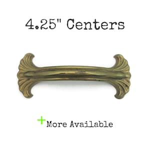 May include: Antique brass-toned cabinet pull with decorative ends. The pull measures 4.25 inches between the centers of the mounting holes. The text "4.25" Centers" is above the pull, and "More Available" is below.