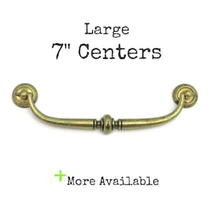 May include: Antique brass drawer pull with a curved handle and circular end plates. The text "Large 7" Centers" is printed above the pull, and "More Available" is printed below. The pull measures 7 inches (17.78 cm) between the center of the mounting holes.