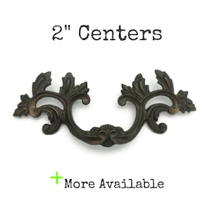 May include: Ornate bronze-colored drawer pull with a decorative floral design. The pull has a 2-inch center-to-center measurement. The text "2" Centers" is above the pull, and "More Available" is below.