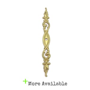 May include: A brass-colored, ornate cabinet pull with a symmetrical design. The pull features a central oval shape with a keyhole, flanked by swirling, decorative elements and leaf-like accents. The text "More Available" is at the bottom.