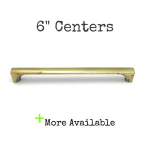 May include: A brass-toned metal cabinet pull with a horizontal bar design and two mounting points. The text 6" Centers is printed above the handle, and a green plus sign with the text More Available is printed below the handle.