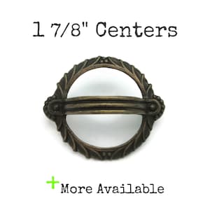 May include: Antique bronze-coloured drawer pull with a decorative oval design. The pull has a ring shape with a horizontal bar across the centre. The text "1 7/8" Centers" is above the pull, and "More Available" is below.
