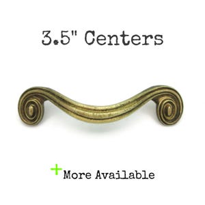 May include: Antique brass-colored cabinet pull with a curved design and scroll ends. The text "3.5" Centers" is above the pull, and "More Available" is below. The pull measures 3.5 inches in length.