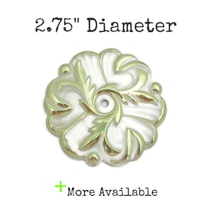 May include: A white and gold decorative rosette with a 2.75 inch diameter. The rosette has a floral design with a central hole.