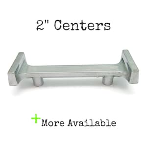 May include: A silver-colored cabinet pull with a 2-inch center-to-center measurement. The pull has a rectangular design with two small cylindrical knobs. The text "2" Centers" and "More Available" are also visible.
