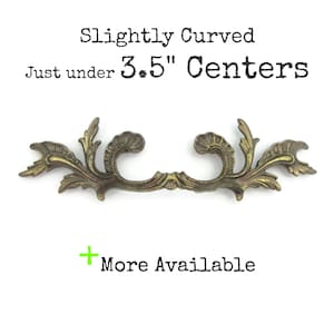 May include: Ornate, antique-style brass drawer pull with a curved design. The text reads "Slightly Curved Just under 3.5" Centers" and "More Available". The hardware has a detailed, floral motif and is suitable for furniture restoration.