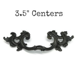 May include: A black metal drawer pull with ornate, scrolling leaf-like details and a curved handle design. The text 3.5" Centers is printed above the hardware, indicating the distance between the two mounting points for this furniture handle.