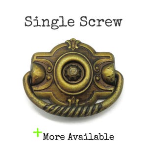 May include: An antique brass-toned metal drawer pull with a rectangular backplate featuring a central circular motif and a curved, twisted drop handle. The text "Single Screw" appears above the hardware, and "+ More Available" is below.
