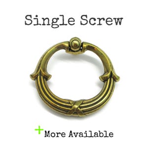 May include: A brass-toned metal ring pull handle with a circular design and a central mounting point. The text "Single Screw" appears above the handle, and the text "+ More Available" is positioned below the handle on a plain white background.
