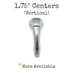 May include: A silver-toned metal hook with a curved, elongated shape and a flat mounting top. The text 1.75" Centers (Vertical) is printed above the hook, and a green plus sign with the text More Available is printed below the hook.