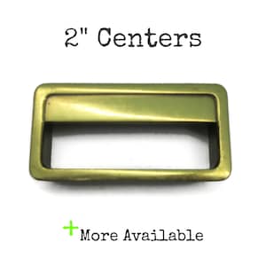 May include: A rectangular brass-toned metal drawer pull with a smooth finish and a central horizontal opening. The text 2" Centers is printed above the hardware, and the text + More Available is printed below the hardware.