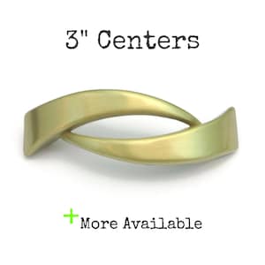 May include: A gold-toned cabinet pull with a curved, flowing design. The text "3" Centers" is above the pull, and "More Available" is below. The pull measures 3 inches.
