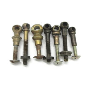 Peut inclure: Un ensemble de sept fixations métalliques anciennes, chacune dotée d'une tête à œillet circulaire et d'une tige filetée. Ces pièces de quincaillerie présentent des nuances variées de laiton vieilli, de bronze et d'acier sombre, avec des écrous.