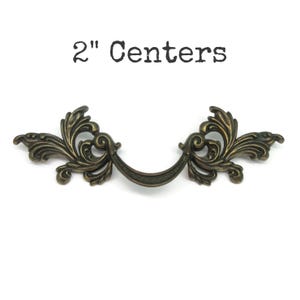 May include: An antique bronze-colored metal drawer pull featuring ornate, symmetrical scrollwork and leaf-like detailing on both sides. The handle has a curved center bar and is labeled with the text 2" Centers above the hardware piece.