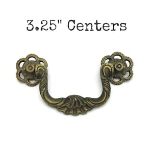 May include: Antique brass drawer pull with a floral design on each end and a curved handle. The text "3.25" Centers" is above the pull. The pull is made of metal and has an aged appearance.