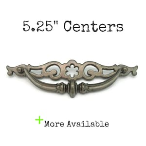 May include: Antique-style drawer pull with a decorative design. The pull is a muted silver color and features a curved handle and ornate detailing. The text "5.25" Centers" is above the pull, and "More Available" is below.