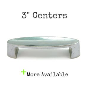 May include: A silver-toned, oval-shaped cabinet pull with a 3" center-to-center measurement. The pull has a raised, flat top and two rectangular supports.