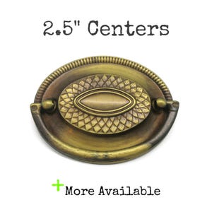 May include: An oval brass drawer pull with a dark antique finish, featuring a central textured leaf pattern and a swinging handle. The text 2.5" Centers and More Available is printed above and below the hardware, indicating the mounting distance.