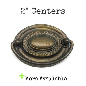 May include: An oval-shaped brass drawer pull with a dark, aged patina finish. The hardware features a central raised oval plate surrounded by a decorative border and a swinging bail handle. The text 2" Centers and More Available is visible.