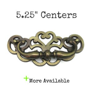 May include: Antique brass drawer pull with an ornate, openwork design. The pull measures 13.3 cm between the centres of the mounting holes. The text "5.25" Centers" is above the pull, and "More Available" is below.
