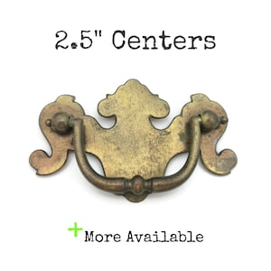 May include: Antique brass drawer pull with a decorative design. The pull has a curved handle and measures 2.5 inches between the centers of the mounting holes. The text "2.5" Centers" is above the pull, and "More Available" is below.