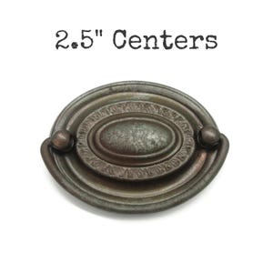 May include: An oval-shaped metal drawer pull with a dark, weathered bronze finish. The hardware features a central oval plate surrounded by a decorative border and a swinging handle. The text 2.5" Centers is printed above the object.