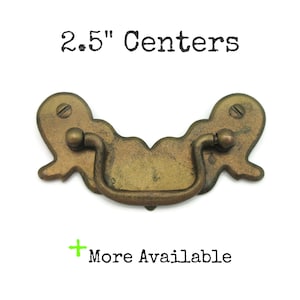 May include: Antique brass drawer pull with a curved handle and decorative ends. The text "2.5" Centers" is printed above the pull. The text "More Available" is printed below the pull.