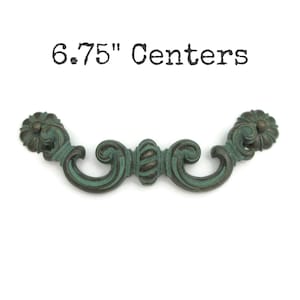May include: A 6.75" center ornate cabinet pull with a green patina finish. The pull has a scrolled design with a decorative ball in the center.