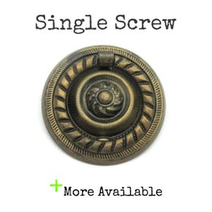 May include: Antique brass-toned drawer pull with a circular design. The pull features a central spiral pattern surrounded by a rope-like border. The text "Single Screw" is above the pull, and "More Available" is below.