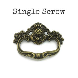 May include: An antique brass-toned drawer pull with an ornate design. The pull features a curved handle and decorative detailing, including a woven pattern in the center. The text "Single Screw" is above the pull.