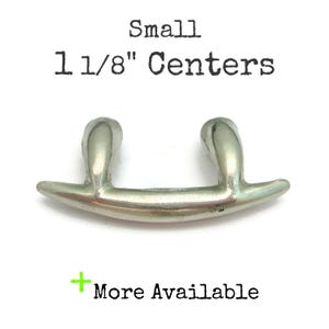 May include: A small, silver-toned cabinet pull with a curved design. The text "Small 1 1/8" Centers" is above the pull, and "+ More Available" is below. The hardware is designed for furniture.