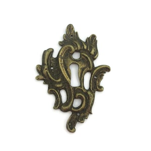 May include: Ornate antique brass keyhole cover with a detailed, symmetrical design. The cover features scrolling leaf and floral patterns, and a keyhole in the center. The metal has a distressed finish, giving it a vintage look. Perfect for furniture restoration.