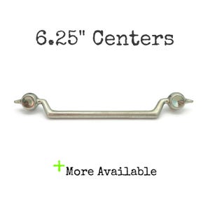 May include: A silver-toned cabinet pull with a 6.25-inch center-to-center measurement. The pull has a straight bar design with decorative ends. The text "6.25" Centers" is above the pull, and "More Available" is below.