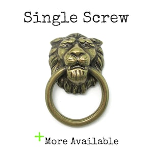 May include: A brass-toned metal lion head door knocker with a circular ring pull. The text "Single Screw" appears at the top, and the text "+ More Available" is printed at the bottom. The lion features detailed mane textures and facial features.