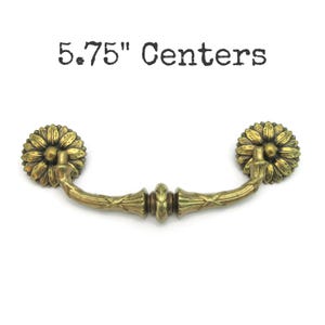 May include: A brass-toned metal drawer pull with a curved handle and two circular floral-shaped backplates. The hardware features a weathered finish and the text 5.75" Centers is printed above the object, indicating the distance between the mounting points.