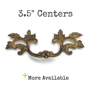 May include: An antique brass-toned metal drawer pull with an ornate, scrolling leaf design. The text 3.5" Centers is printed above the hardware, and a green plus sign with the text More Available is printed below the object.